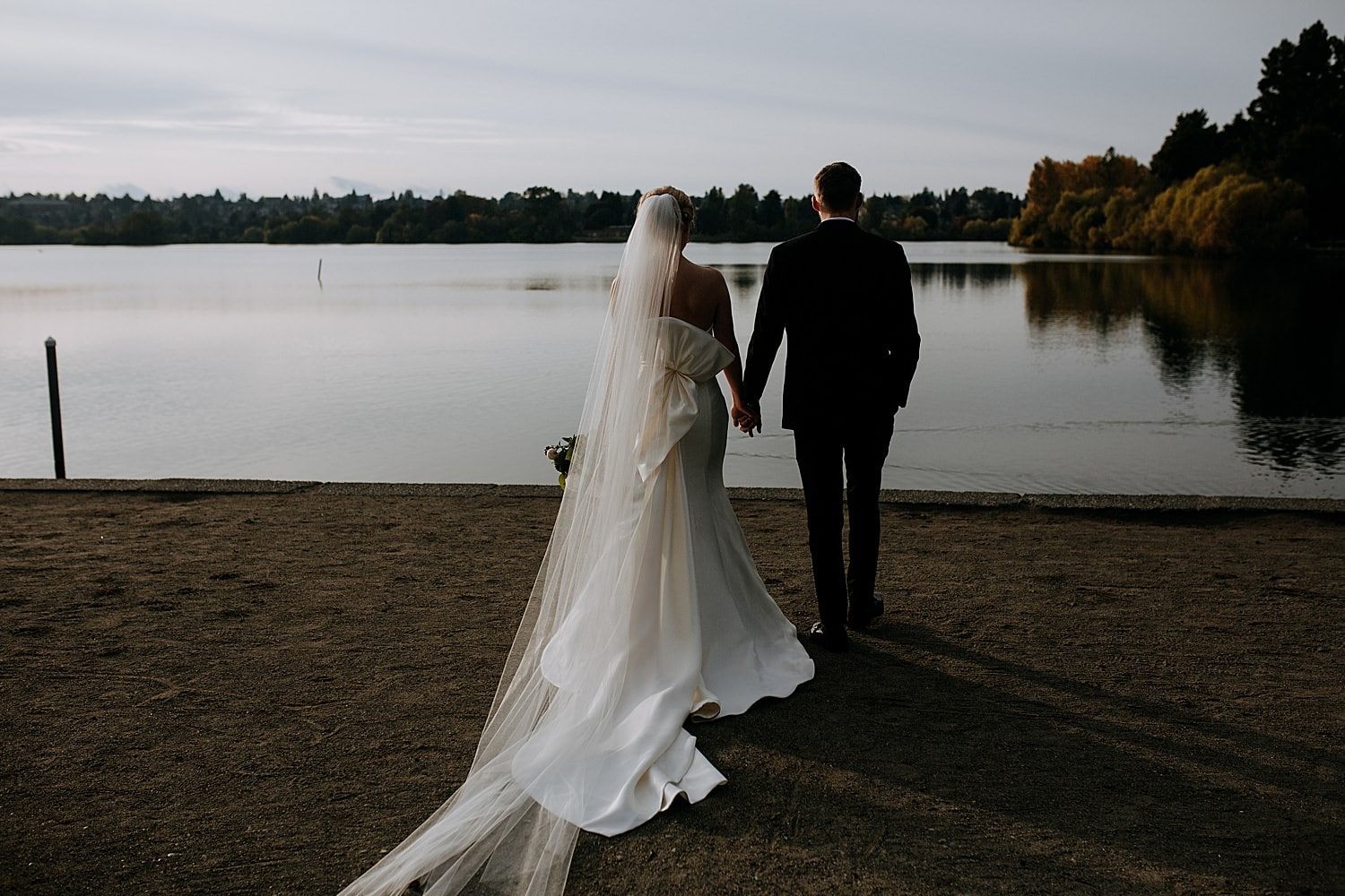 dark and moody portrait of the bride and groom walking away towards Green Lake in Seattle 415 Westlake Wedding by Seattle Wedding Photographer Marcela Pulido