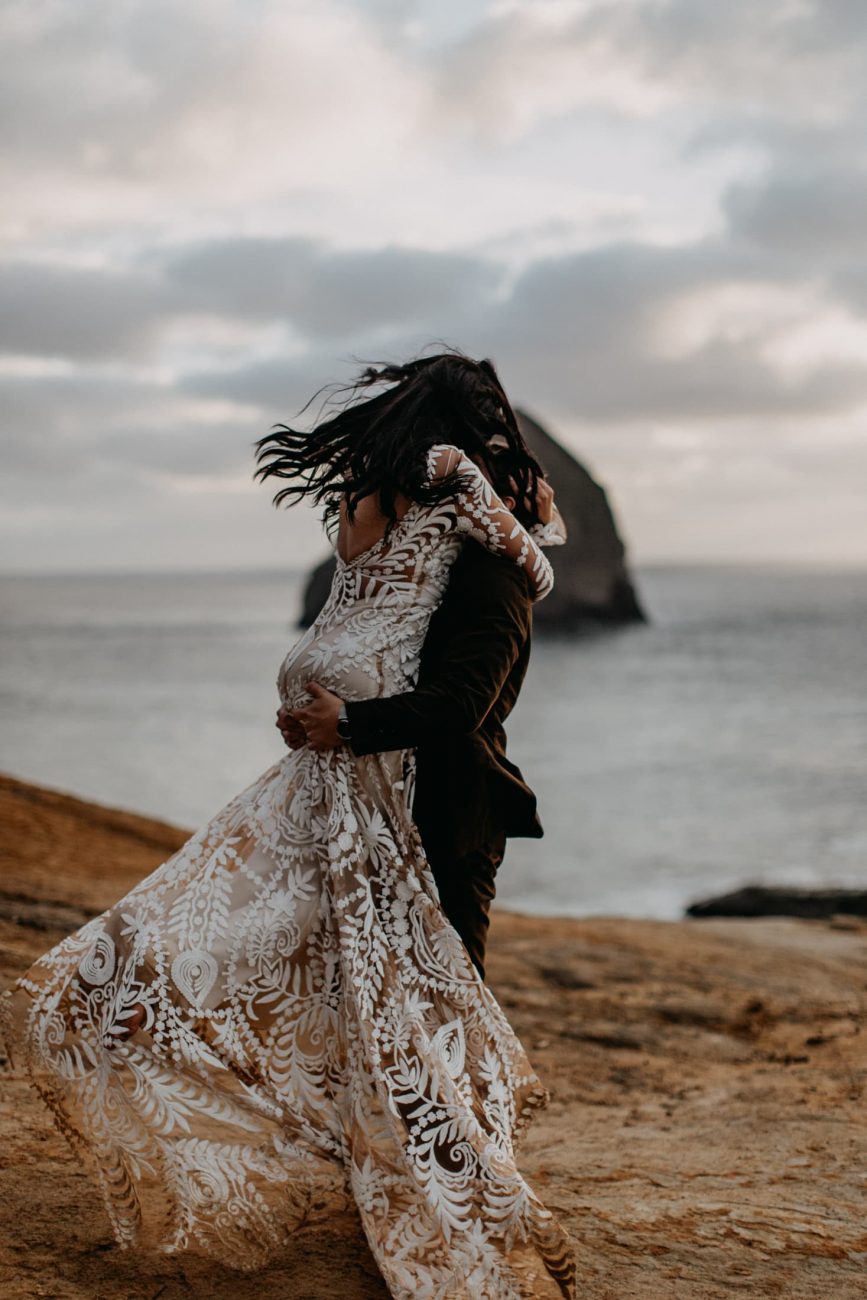 romantic elopement photo of groom picking his bride up and spinning her around wearing a gorgeous rue de seine wedding gown at cape kiwanda photographed by marcela pulido photography portland wedding photographer