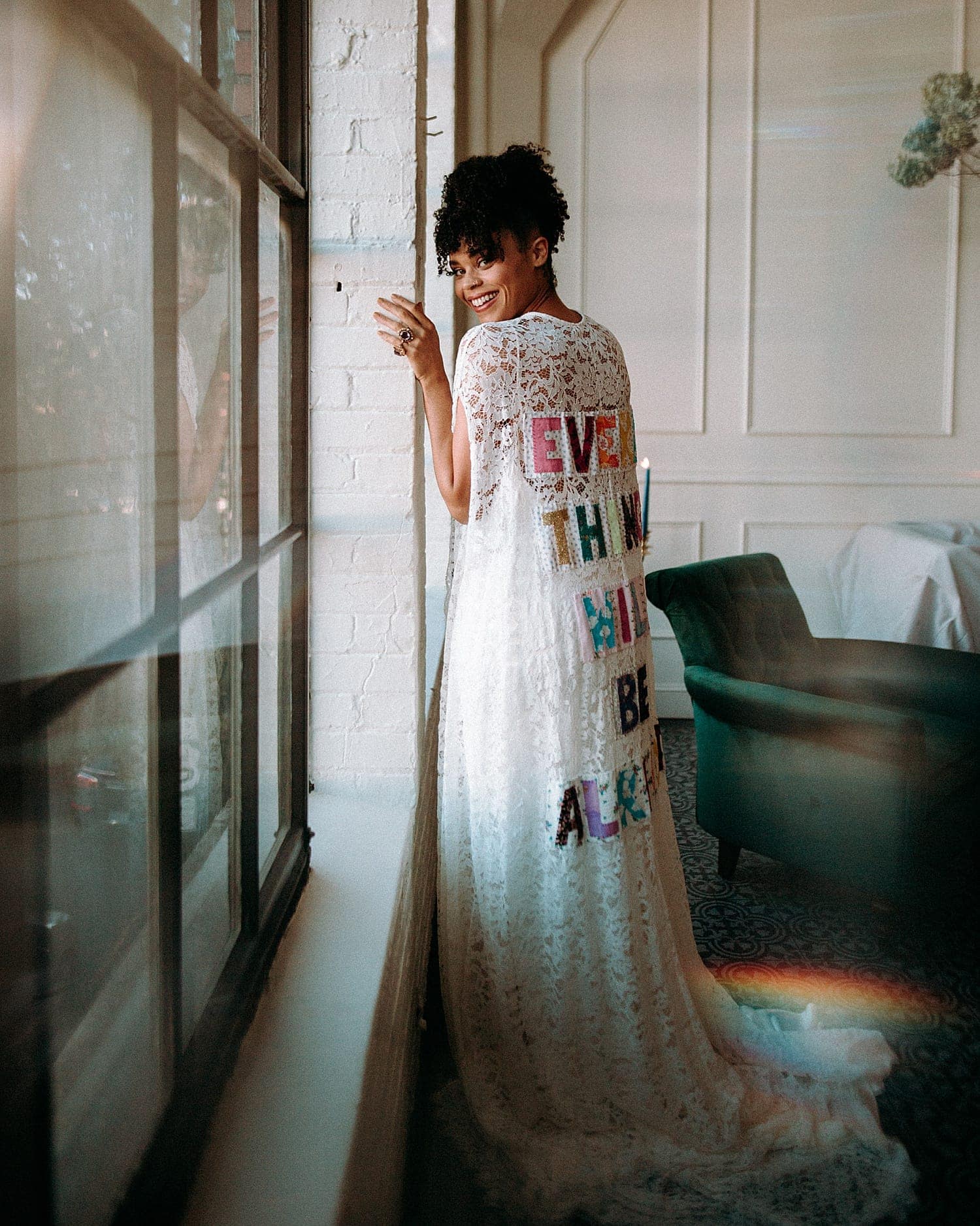 dreamy and ethereal portrait of a black bride with hazel eyes and curly hair grinning leaning up against a window wearing a lace cape by Claire La Faye that says "every thing will be alright"