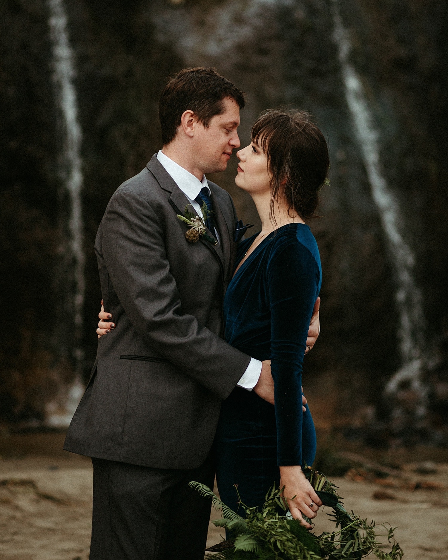 newlywed couple embracing in front of waterfall wearing dark blue velvet wedding dress at this oregon coast hug point elopement captured by marcela pulido photography portland oregon wedding and elopement photographer