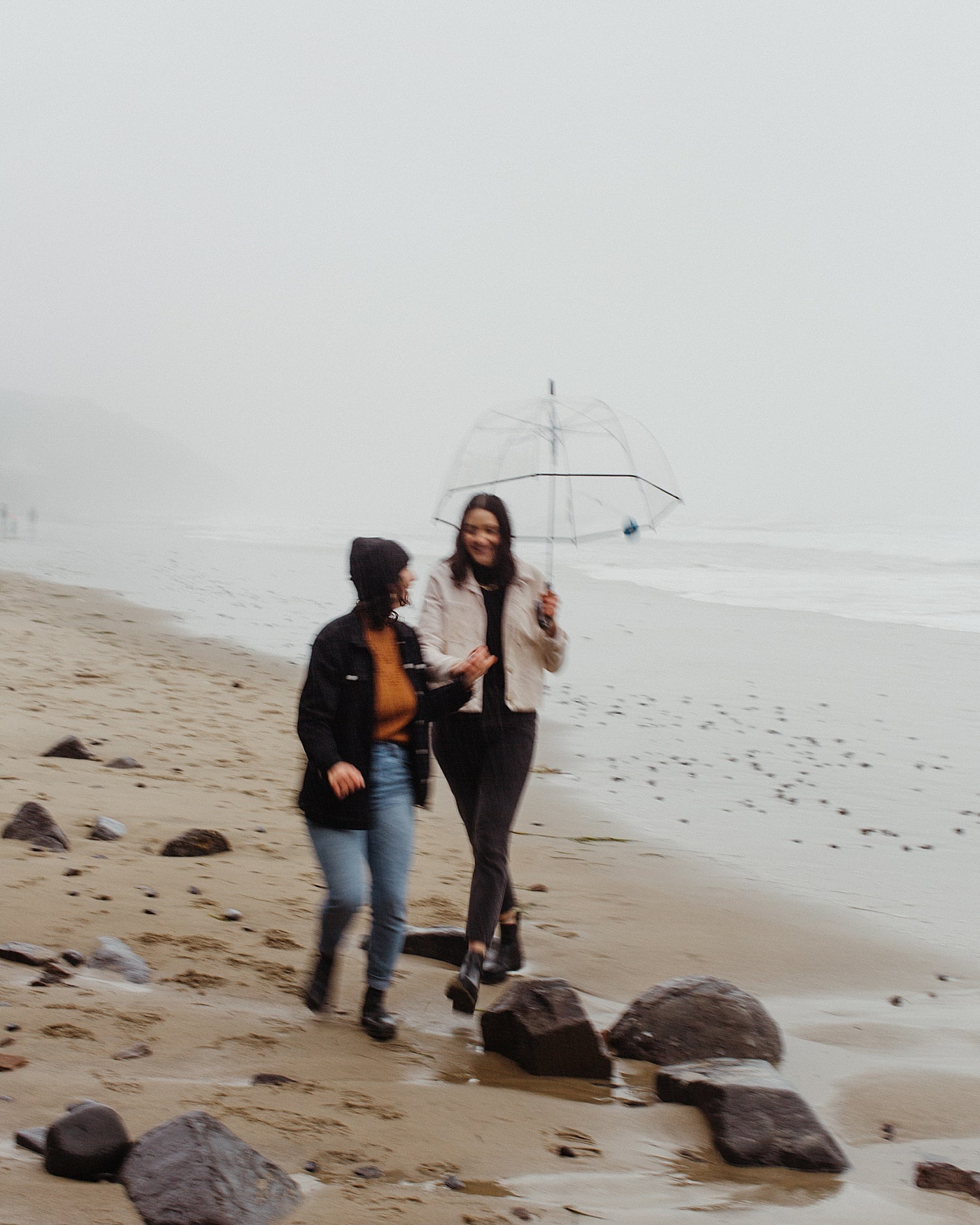 ambient blurry movement shot of same sex lesbian lgbtq+ couple walking on a rainy beach with a clear umbrella by Marcela Pulido Portland Wedding Photographer